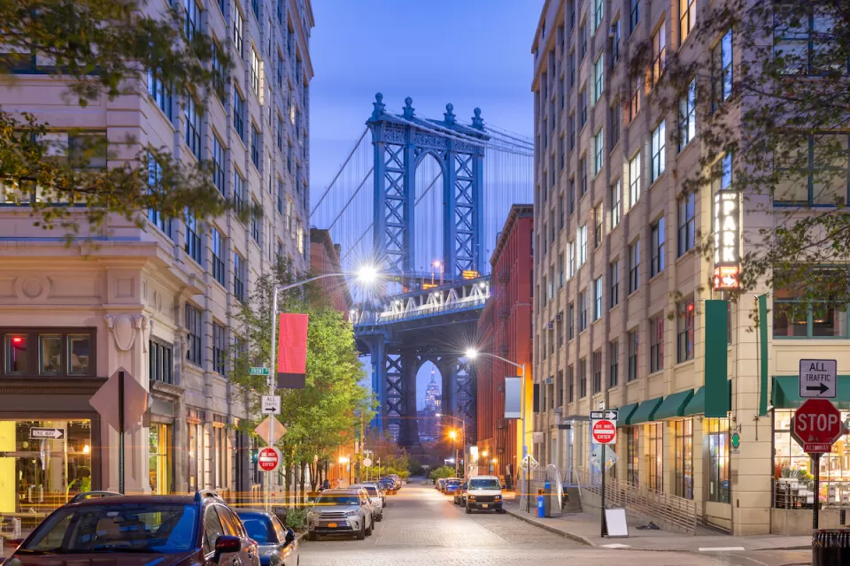 View of Brooklyn Bridge at night from Brooklyn alley with multi-story buildings and parked cars.